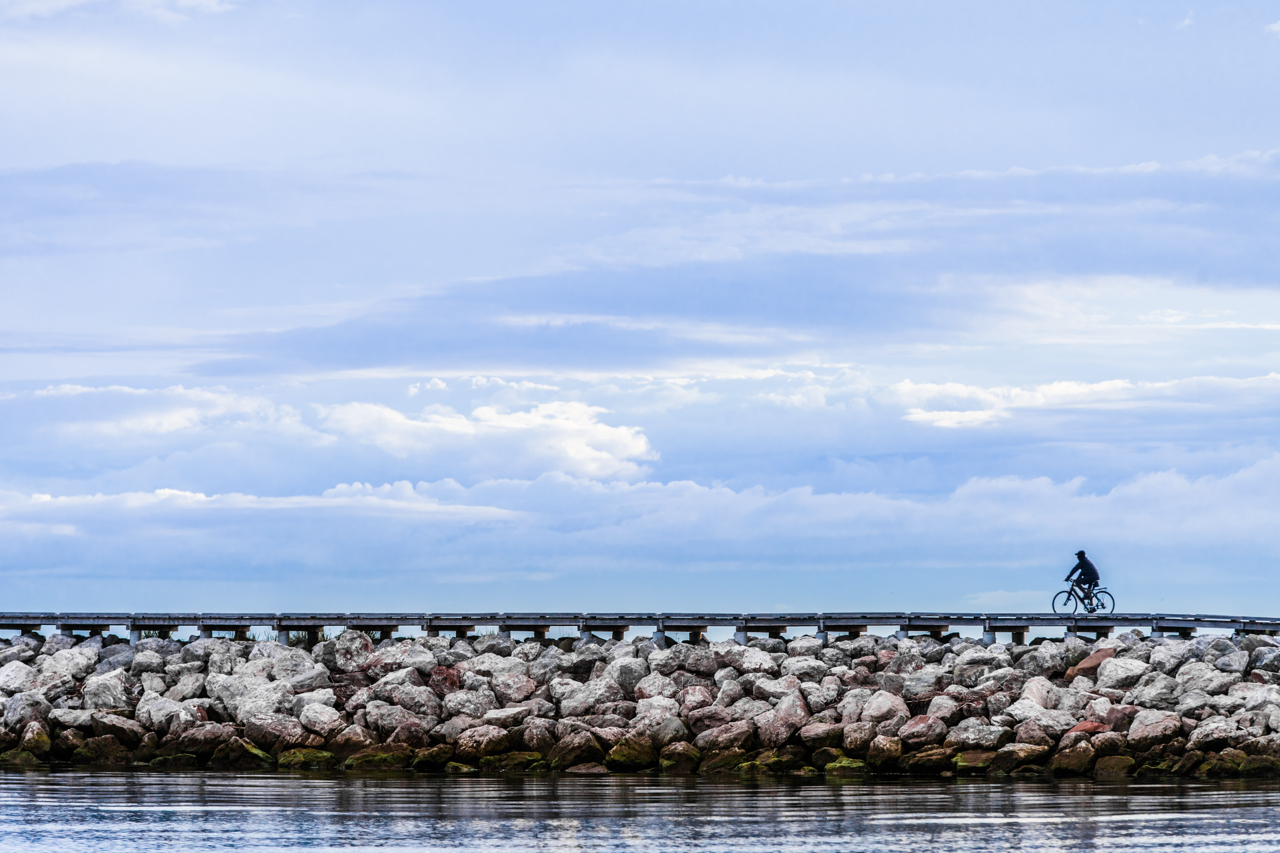 Persona andando en bicicleta sobre un muelle hecho de grandes rocas bajo un cielo azul nublado.